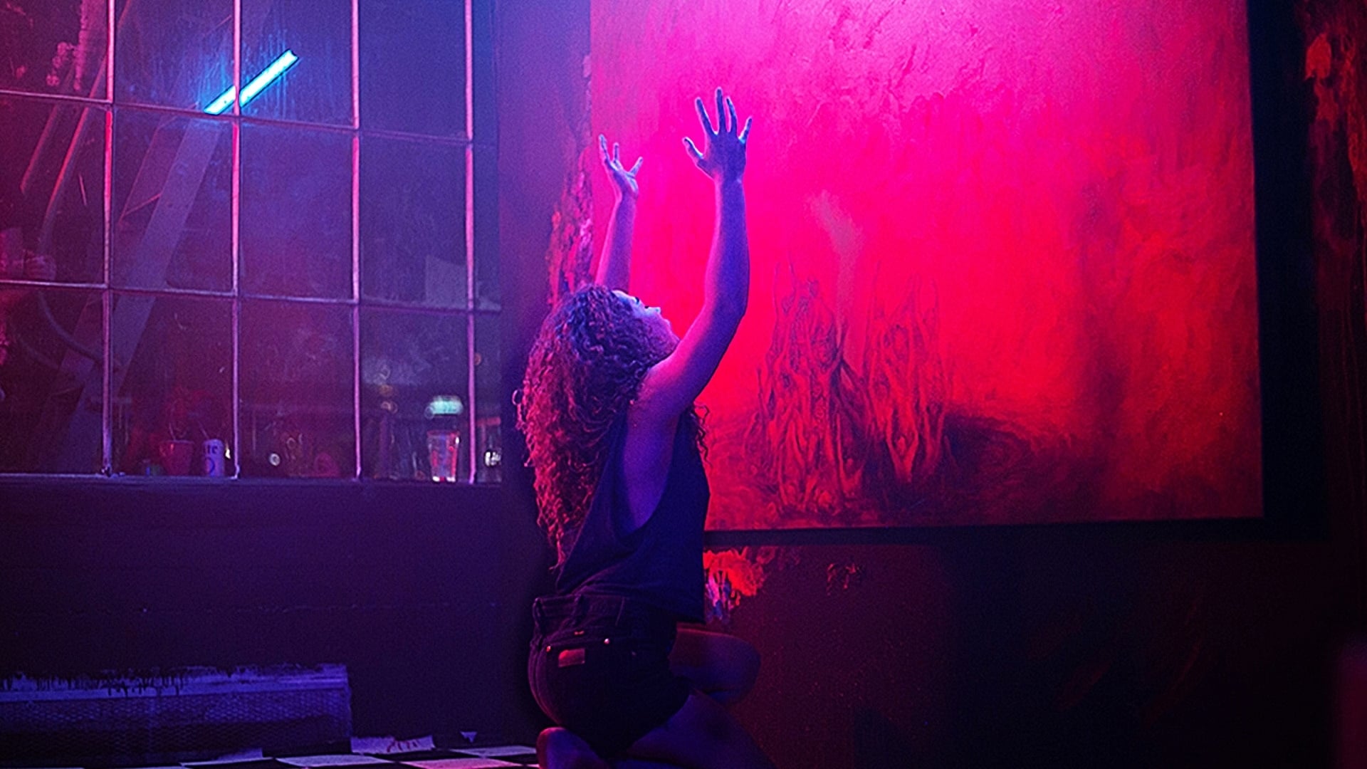 Screenshot from the film Bliss. A woman kneels with her arms raised in front of a painting. The lighting is bright pink and purple.