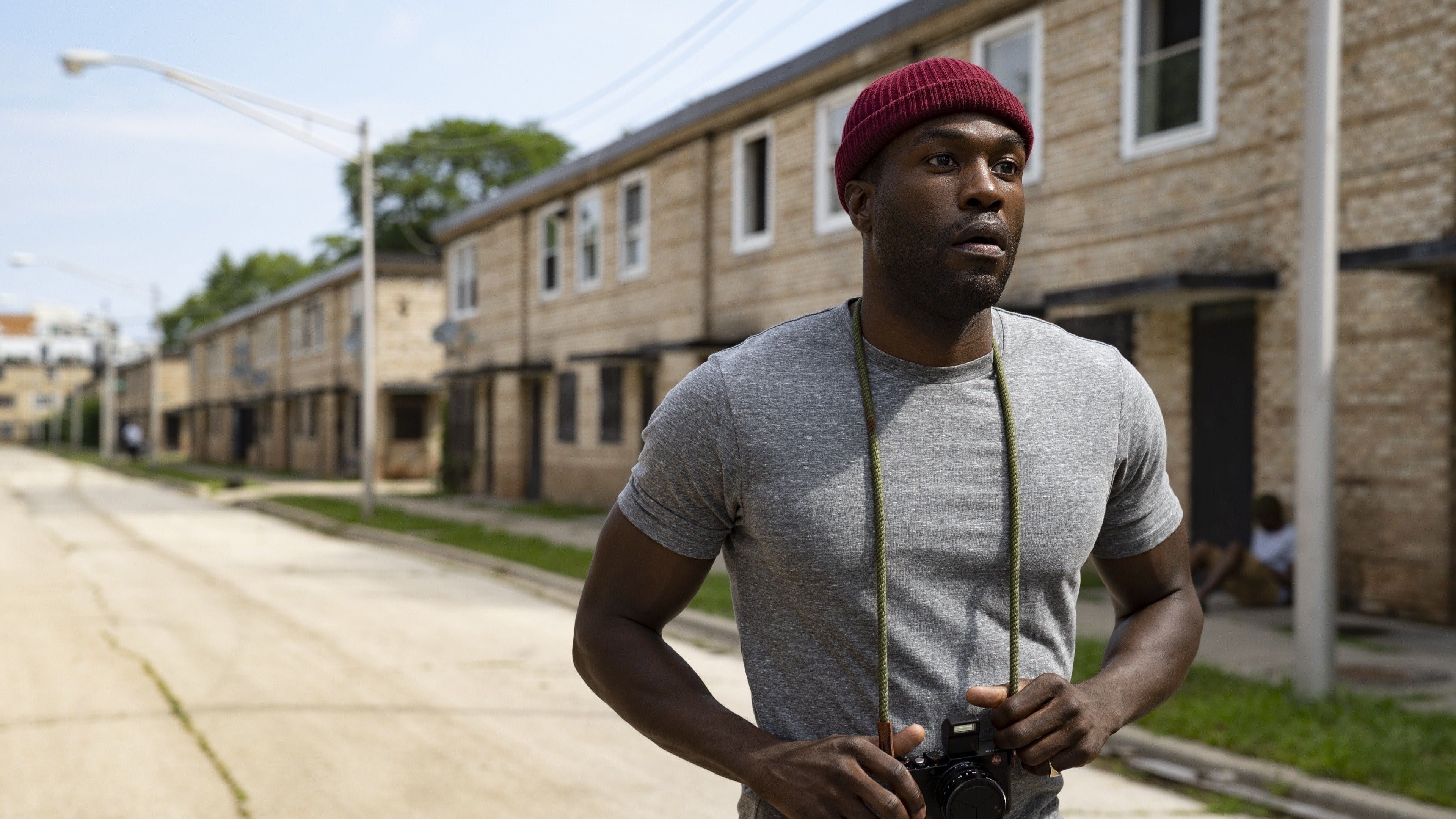 Screenshot from the 2021 film Candyman. A young black man stands on an empty street in the projects with a camera around his neck. 