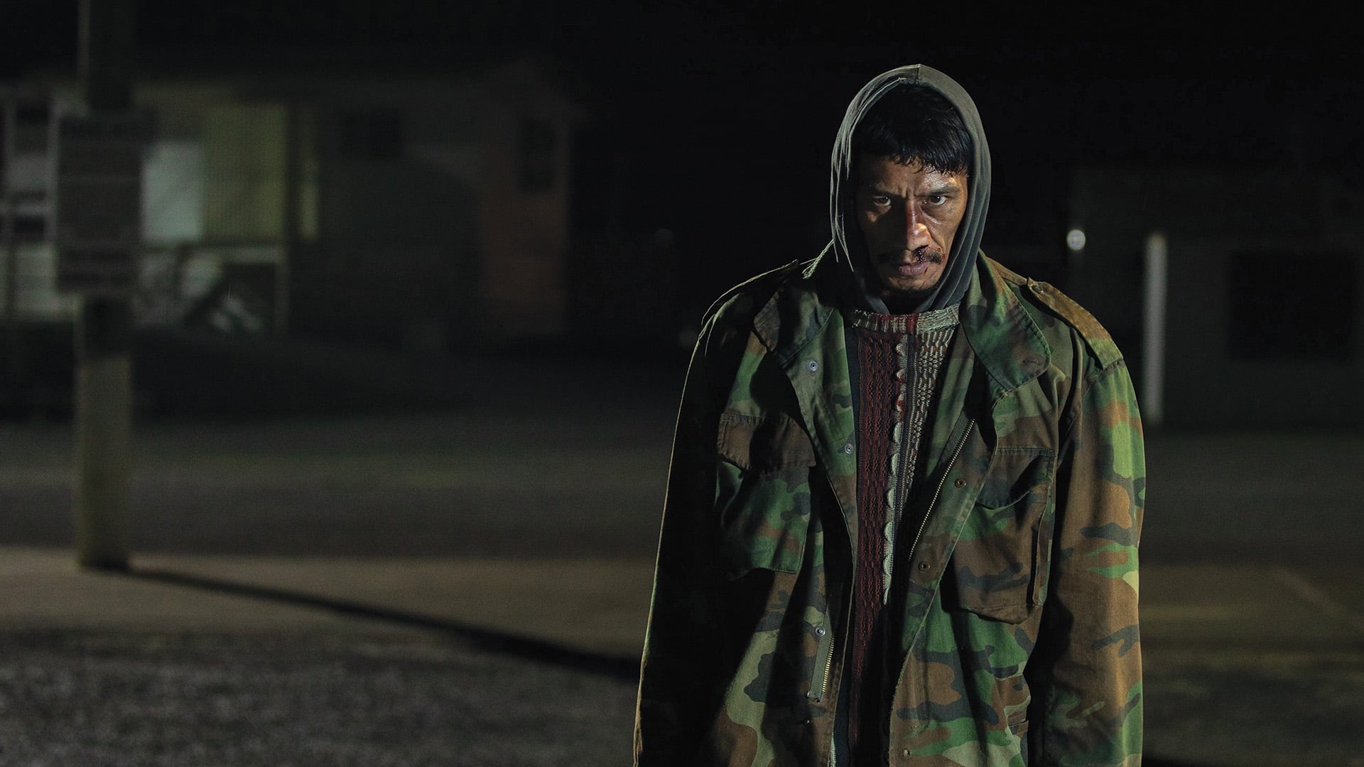 Screenshot from the film Coming Home in the Dark. A man with a bloody face and camo jacket stands and stares menacingly in the dark. 
