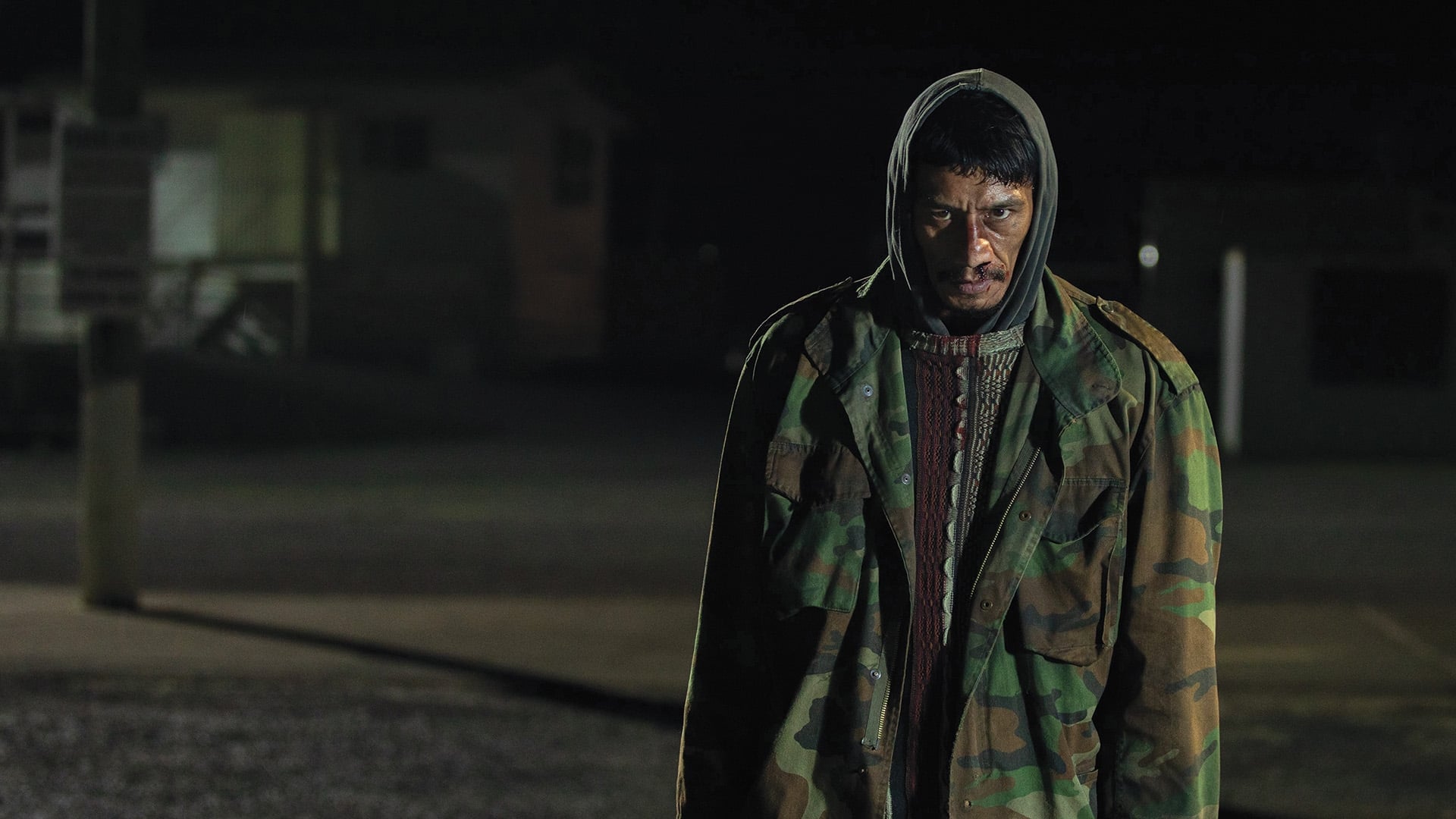 Screenshot from the film Coming Home in the Dark. A man with a bloody face and camo jacket stands and stares menacingly in the dark. 