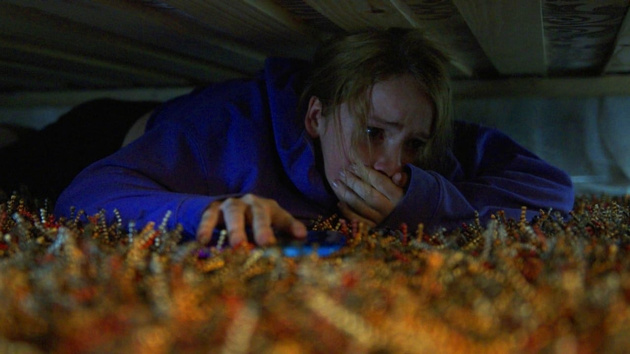 Screenshot from the film Countdown. A woman in a blue jacket hides under the bed covering her mouth. 