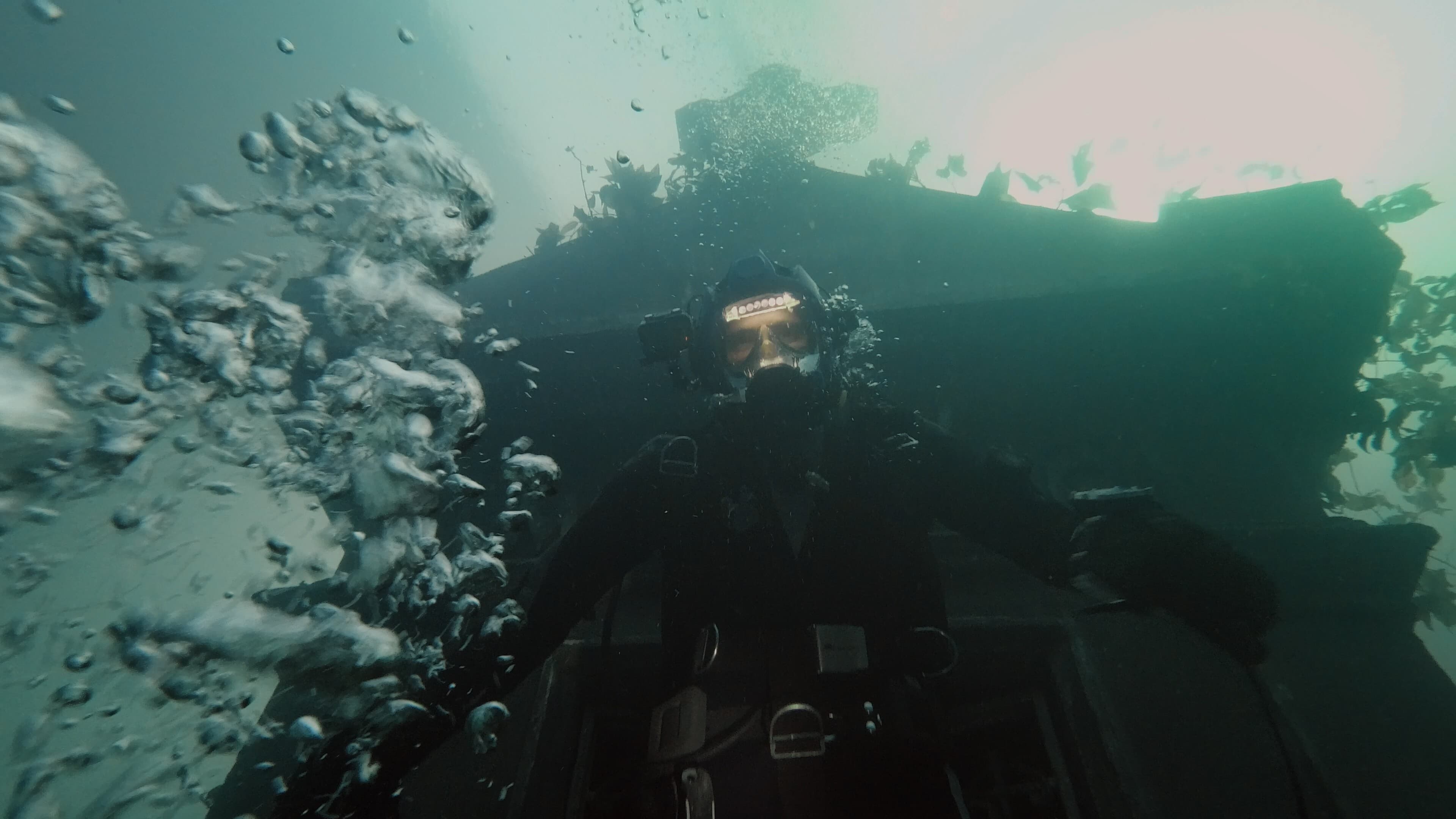 Screenshot from the film The Deep House. A scuba diver looks down with a looming house behind him under water.