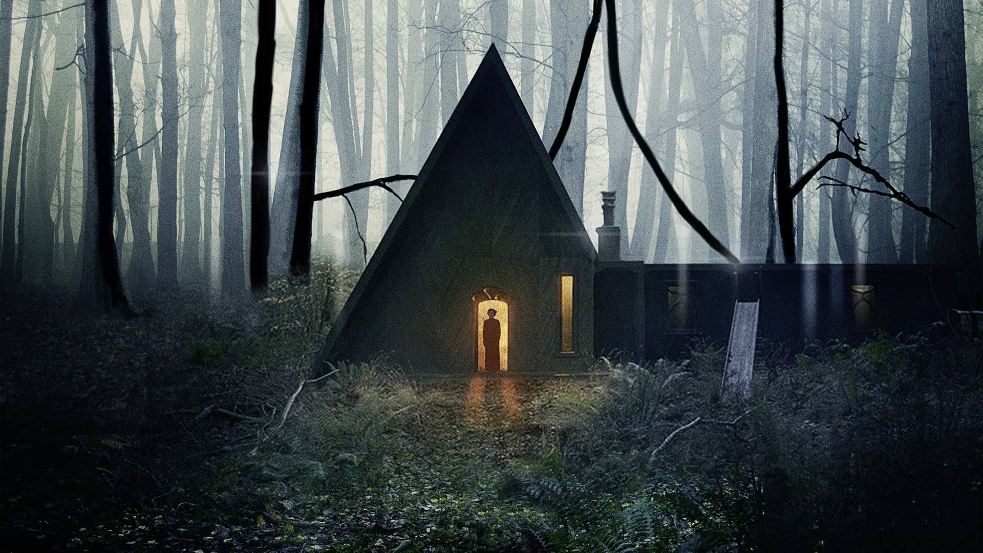 Screenshot from the film Gretel & Hansel. An A-frame cabin sits surrounded by dark woods. The cabin door is open with warm light spilling out while a dark figure of a woman stands in the doorway. 