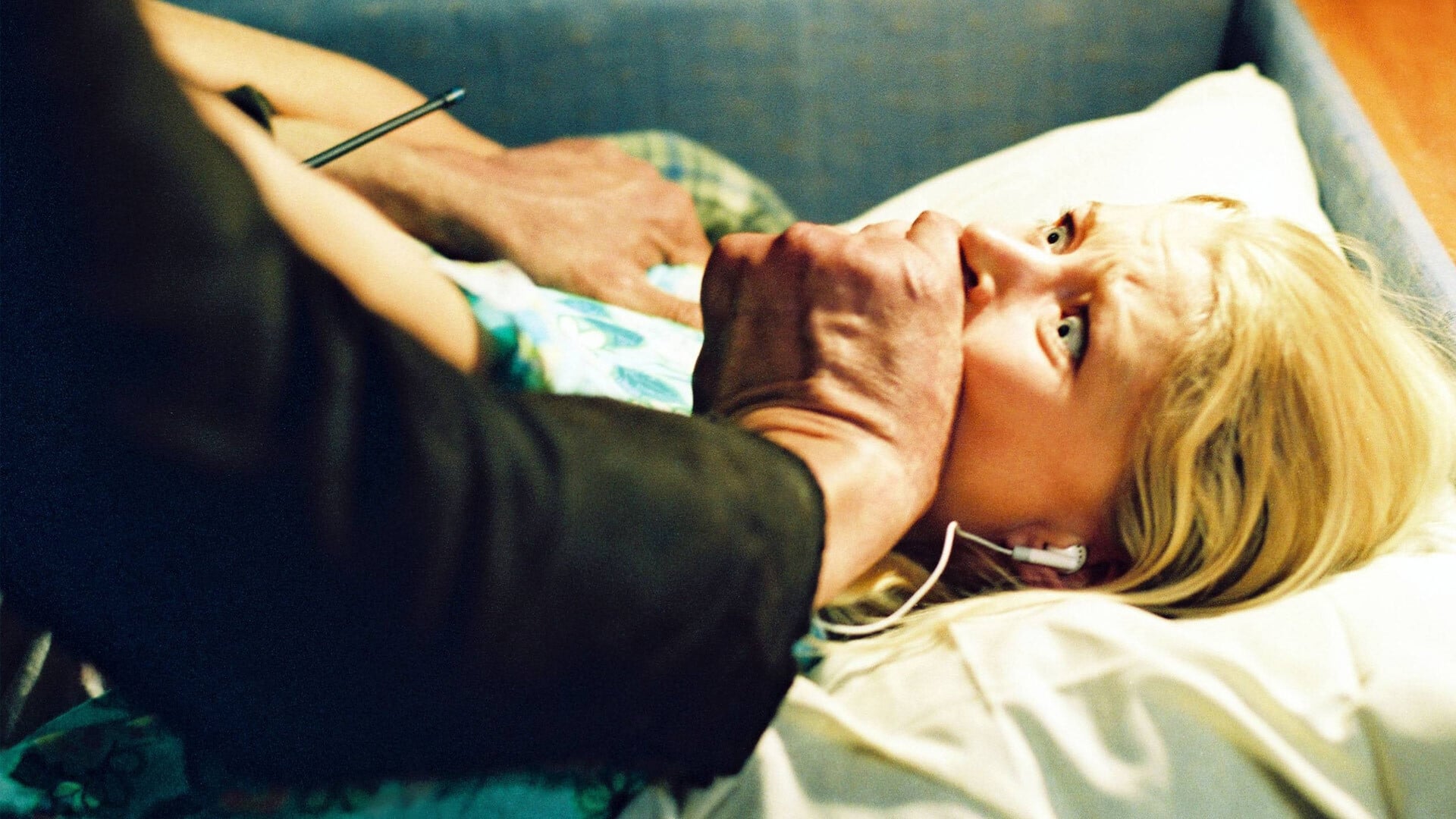 Screenshot from the 2006 film The Hills Have Eyes. A blonde woman is laying in bed with a man covering her mouth. The expression on the woman's face is of terror. 