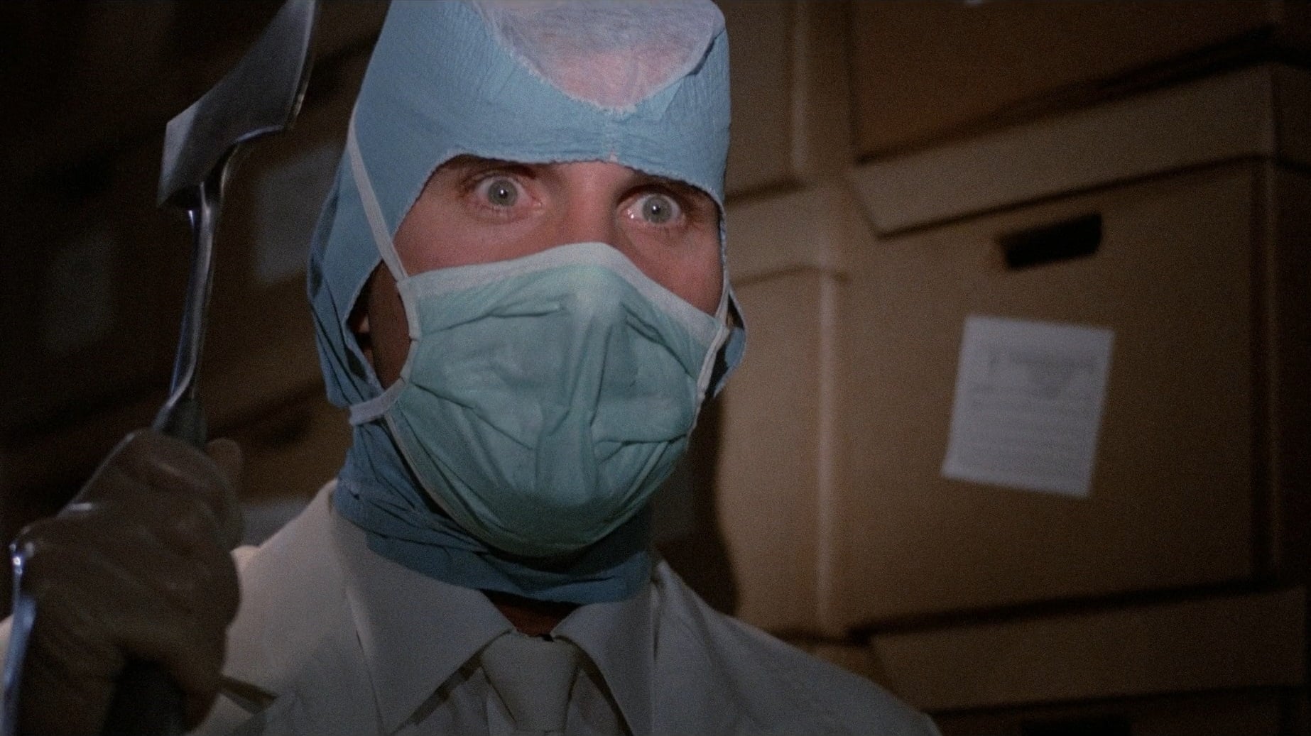 Screenshot from the film Hospital Massacre. A man wearing a doctor's uniform and surgical mask is holding a small axe. 
