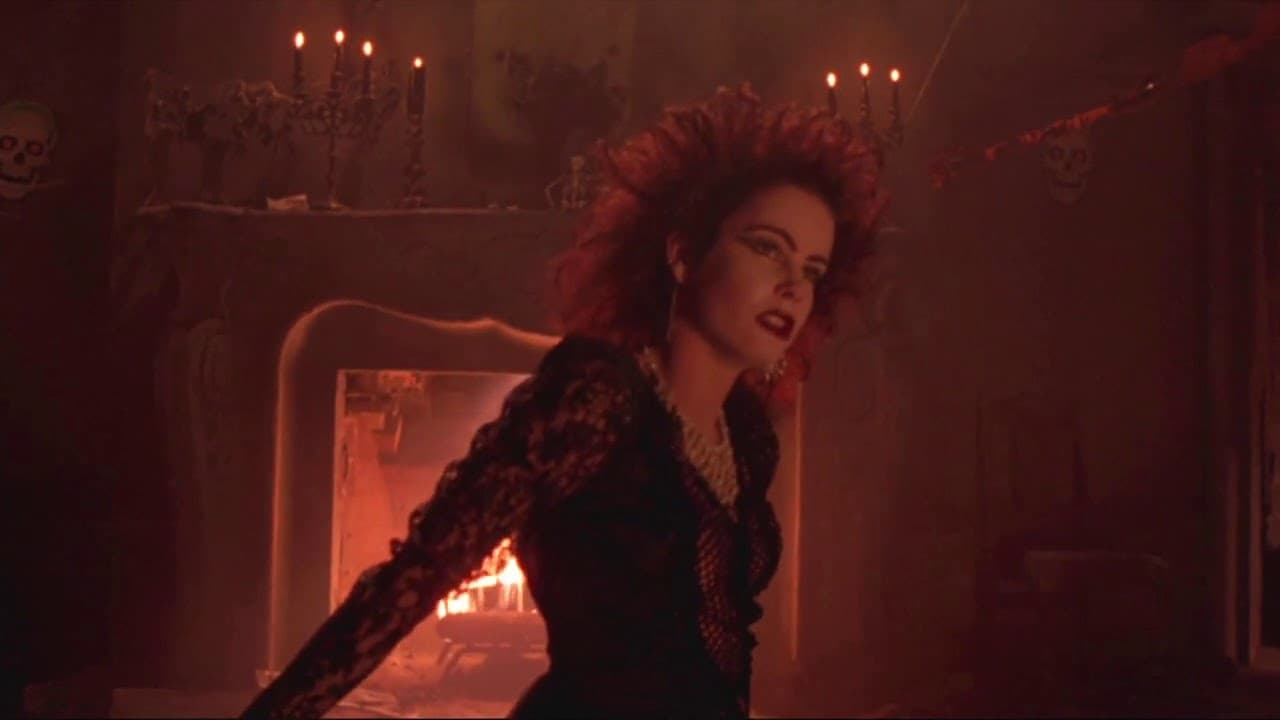 Screenshot from the film Night of the Demons. A young goth woman sits in a black dress in a mansion decorated for Halloween with a fireplace. 