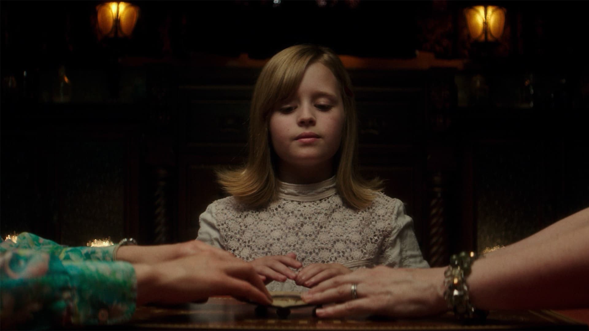 Screenshot from the film Ouija: Origin of Evil. A young girl in a white dress places her hand on a planchet along with two other hands.