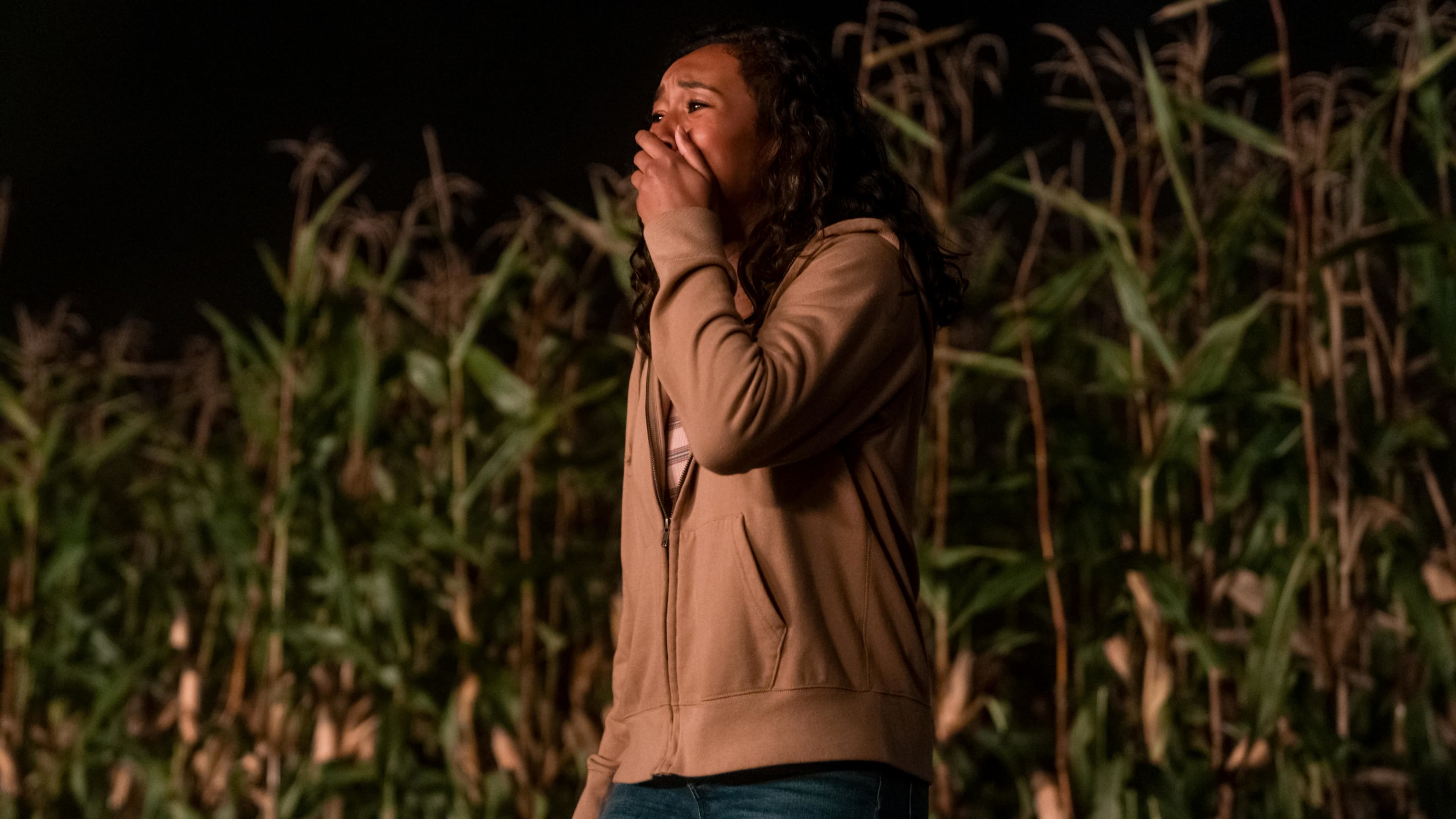 Screenshot from the film There's Someone Inside Your House. A scared girl stands in front of a corn field with her mouth covered in terror. 